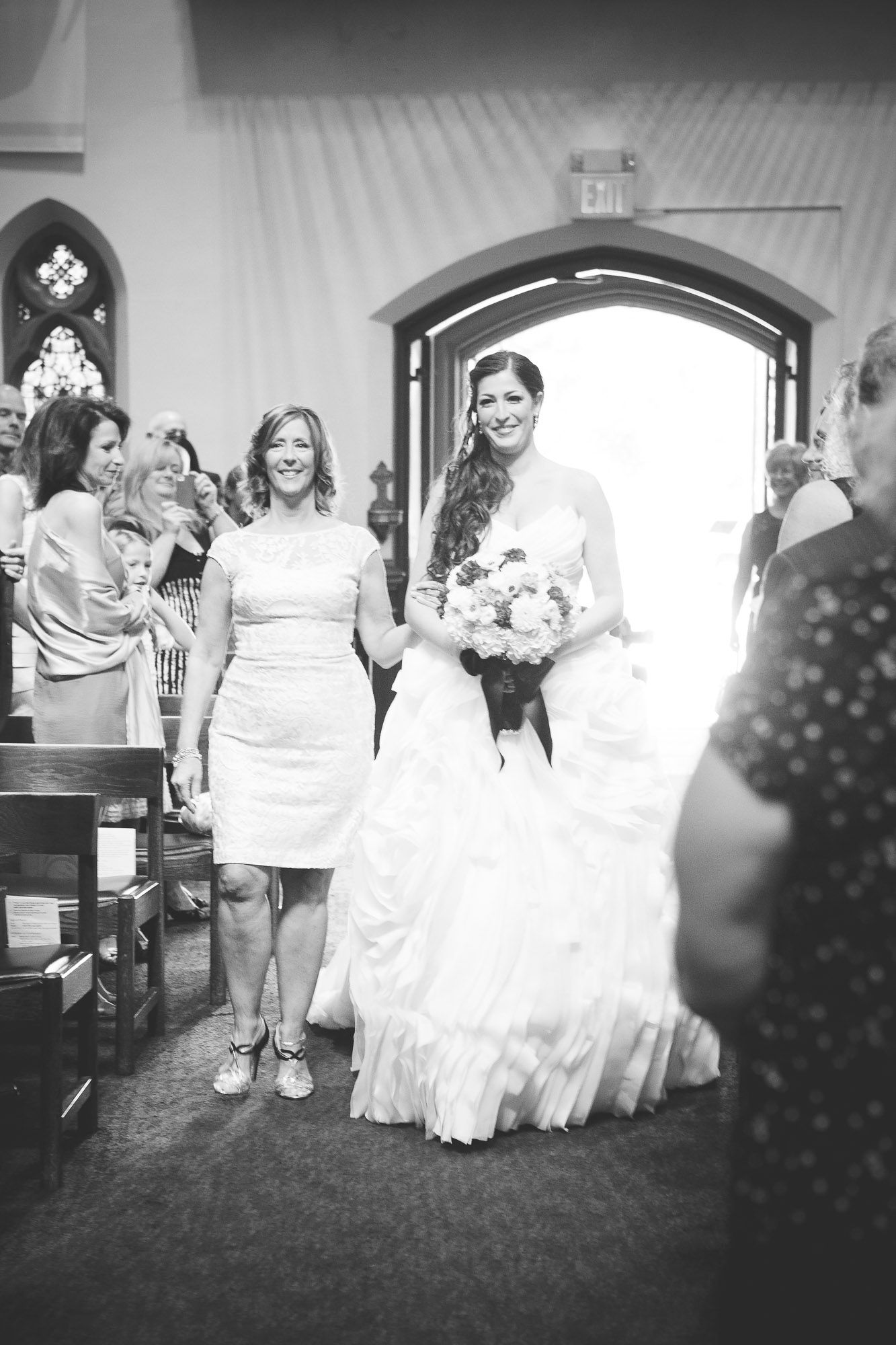 Bride in a ruffled white gown walking down the church aisle with a smiling woman in black and white photo.