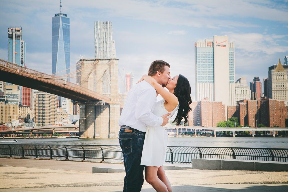 Couple kissing near Brooklyn Bridge with Manhattan skyline and One World Trade Center in background.