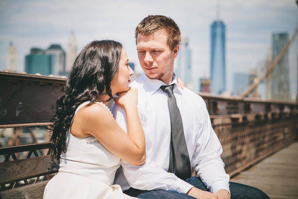 Intimate couple moment on Brooklyn Bridge bench, woman touching mans face with city skyline in background.