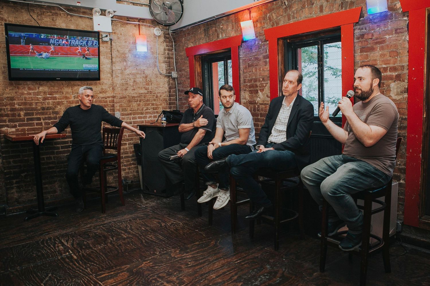 Five men seated on stools in a brick-walled bar during a panel discussion.