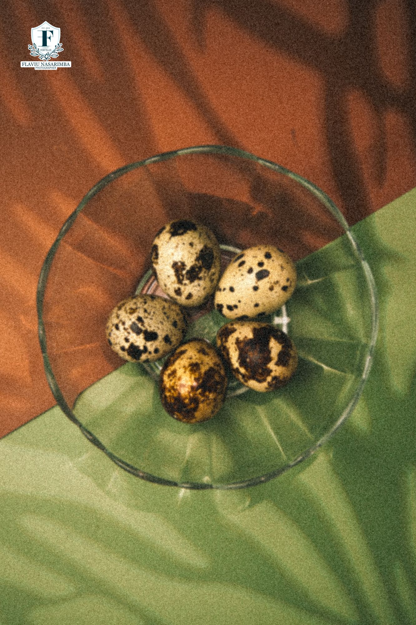 Five speckled quail eggs in a clear glass bowl on green and brown fabric background.