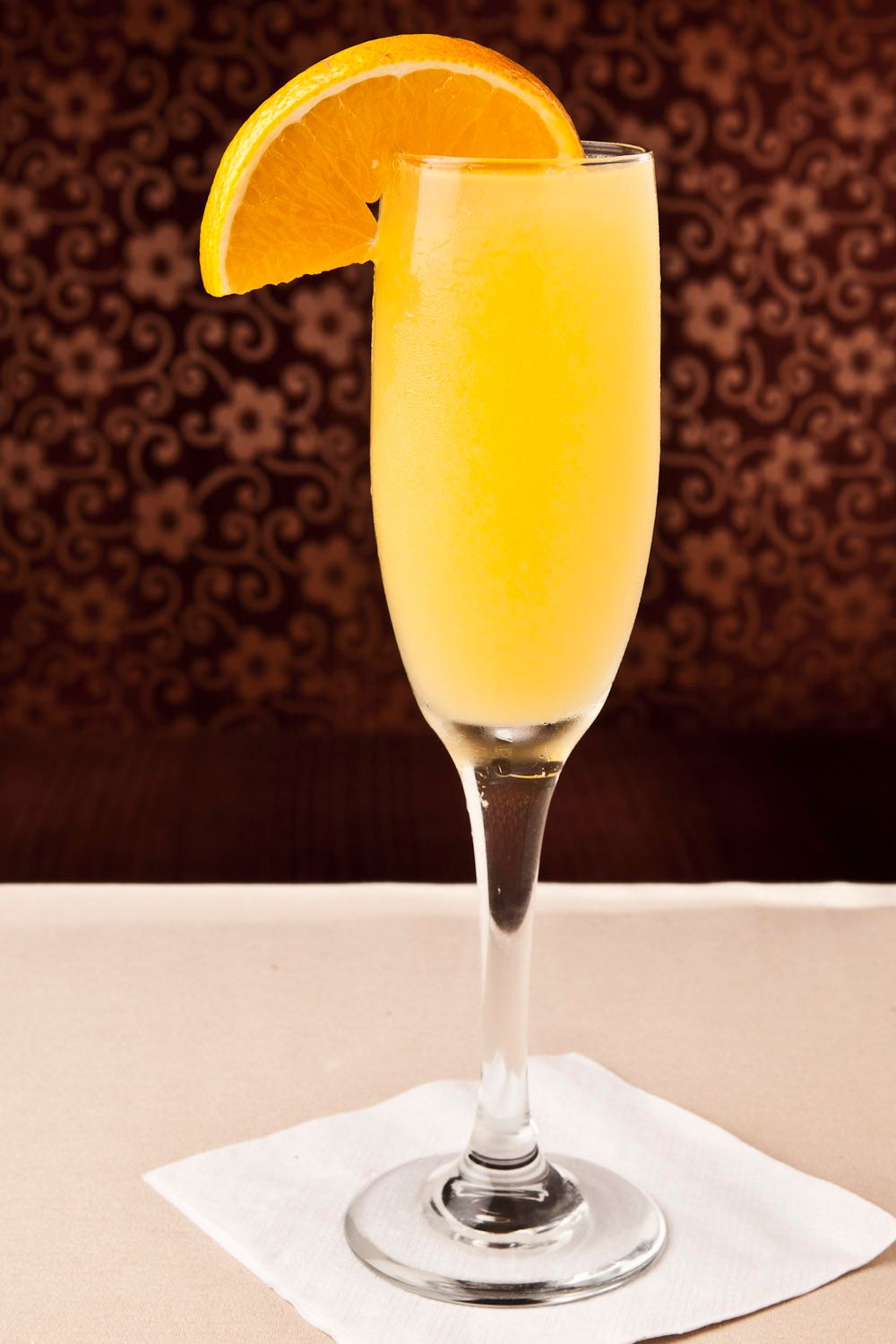 A classic mimosa cocktail in a champagne flute garnished with a fresh orange slice on a white napkin.