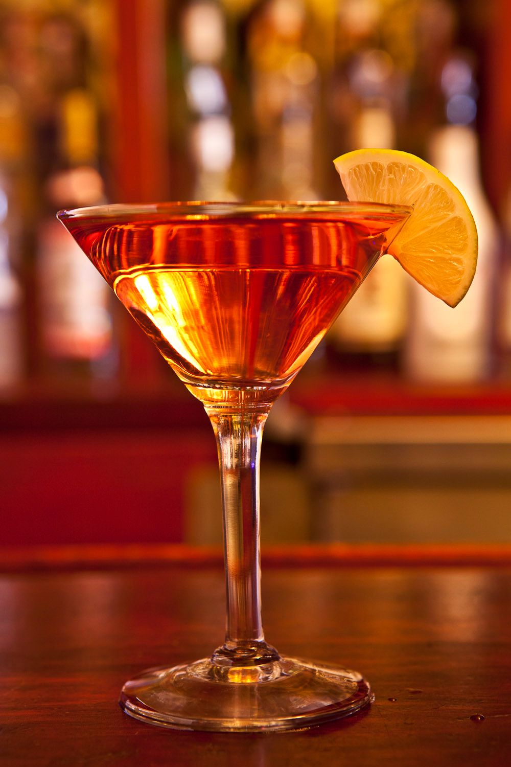 Amber cocktail in a martini glass garnished with an orange slice at a bar.