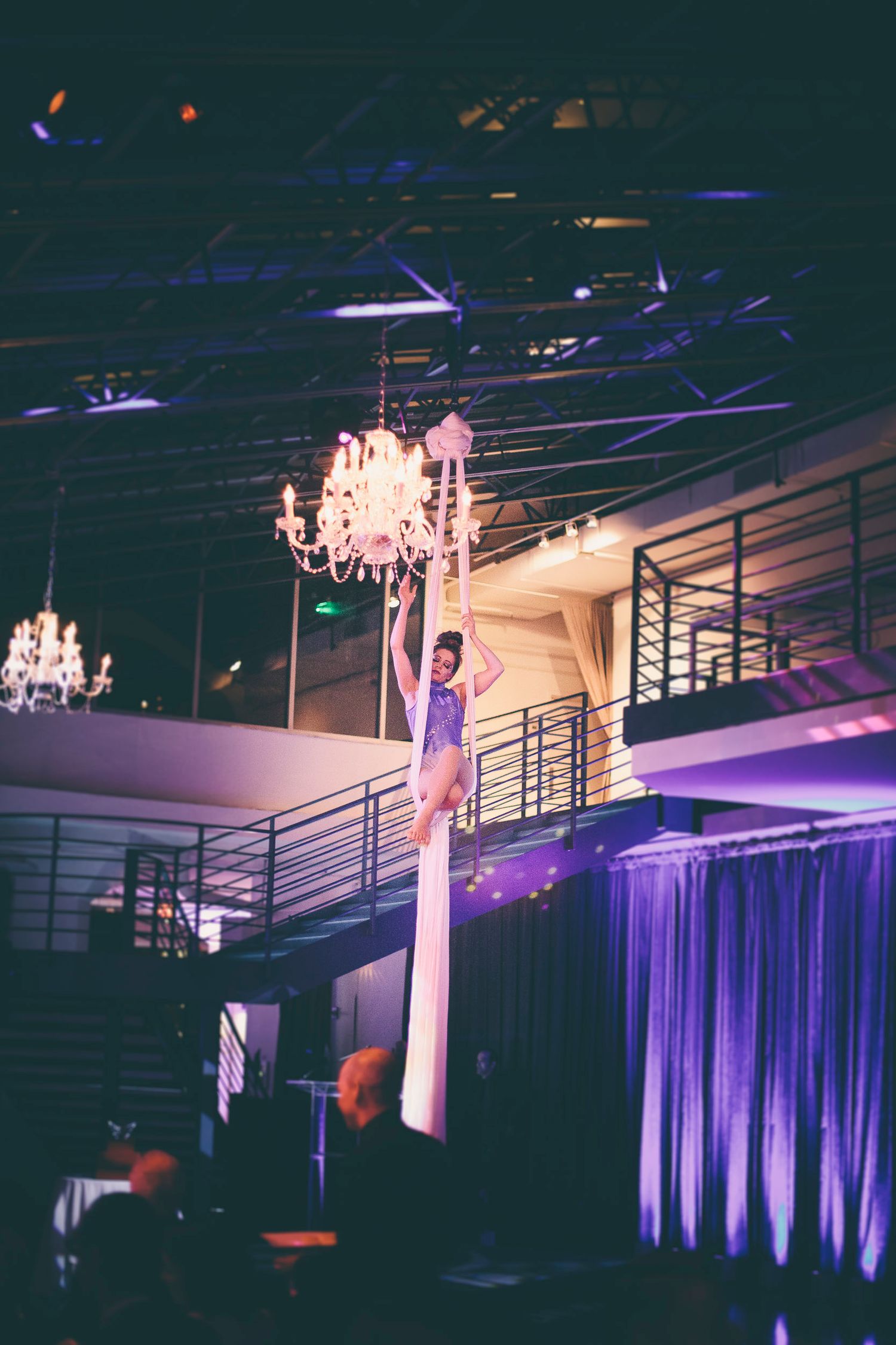 An aerial silk performer climbs high near a chandelier in a dramatic event space with purple lighting.