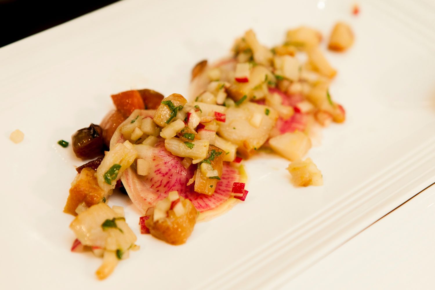 Fine dining appetizer with seared fish, diced vegetable salsa, and red accents on a white plate.