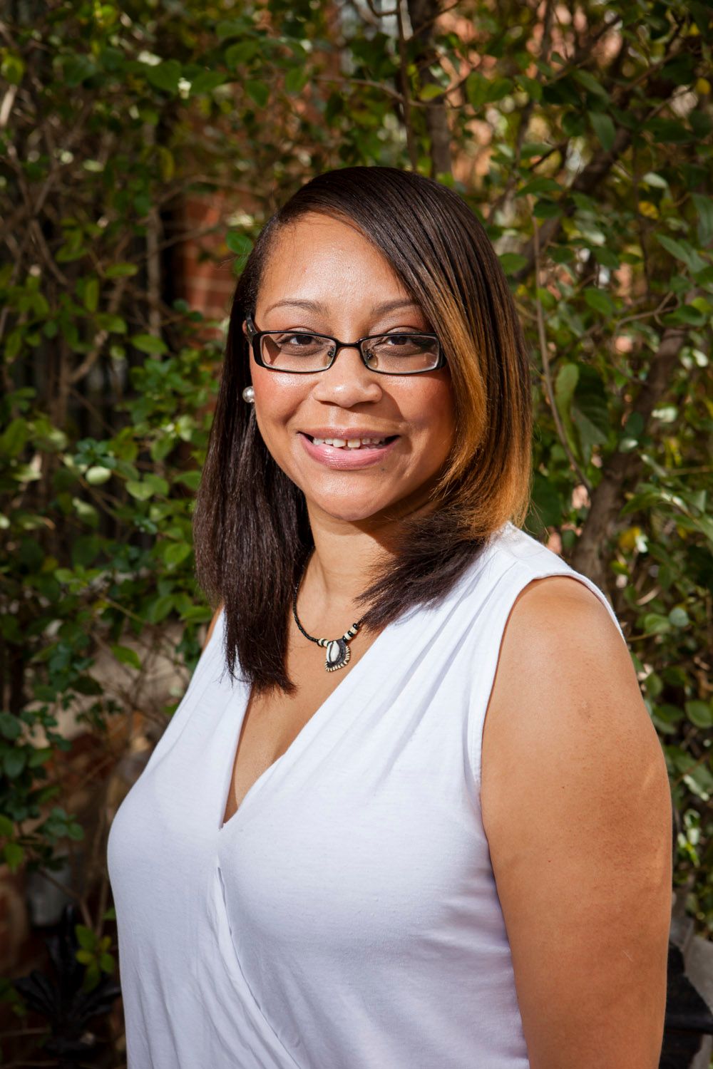 Smiling woman with glasses and white sleeveless top posing outdoors near green foliage.