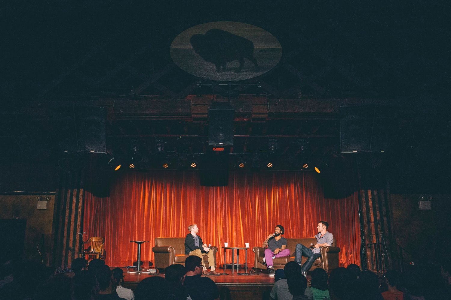 Three speakers on stage under bison logo projection in darkened theater venue.
