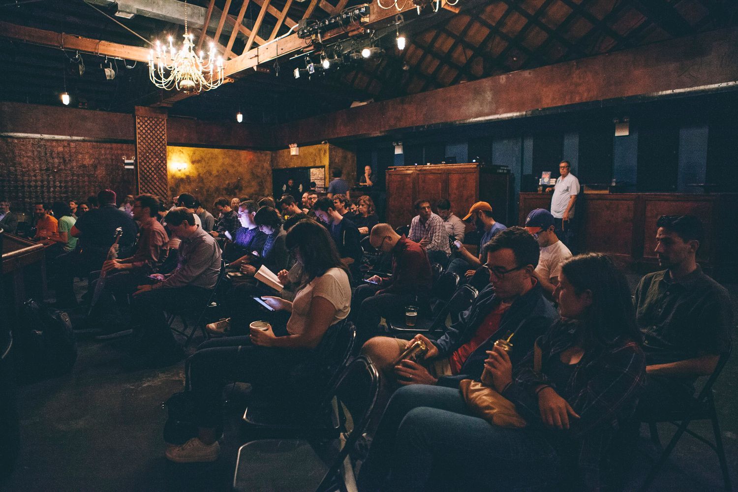 Large audience seated in a rustic venue with chandeliers listening to a speaker on stage.