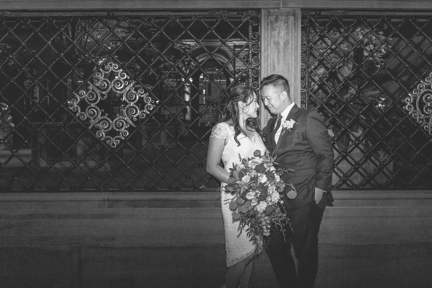 Couple leans close together in black and white photo against decorative iron lattice backdrop.
