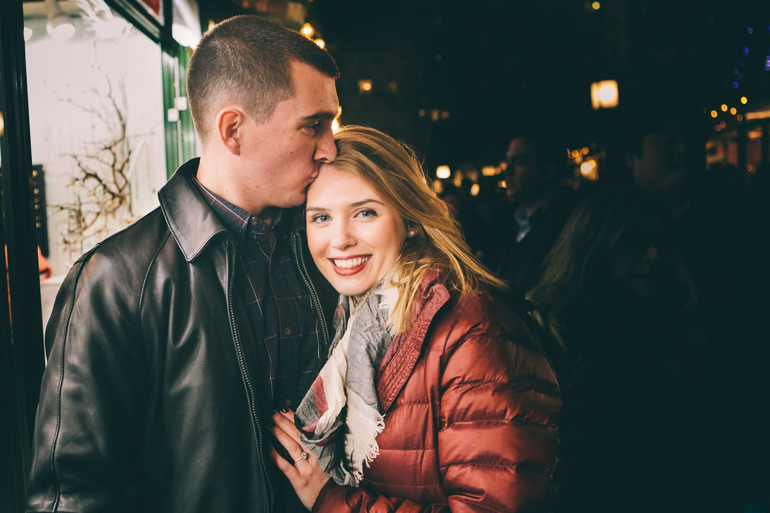 Young man kisses smiling blonde woman on cheek during romantic nighttime holiday stroll.