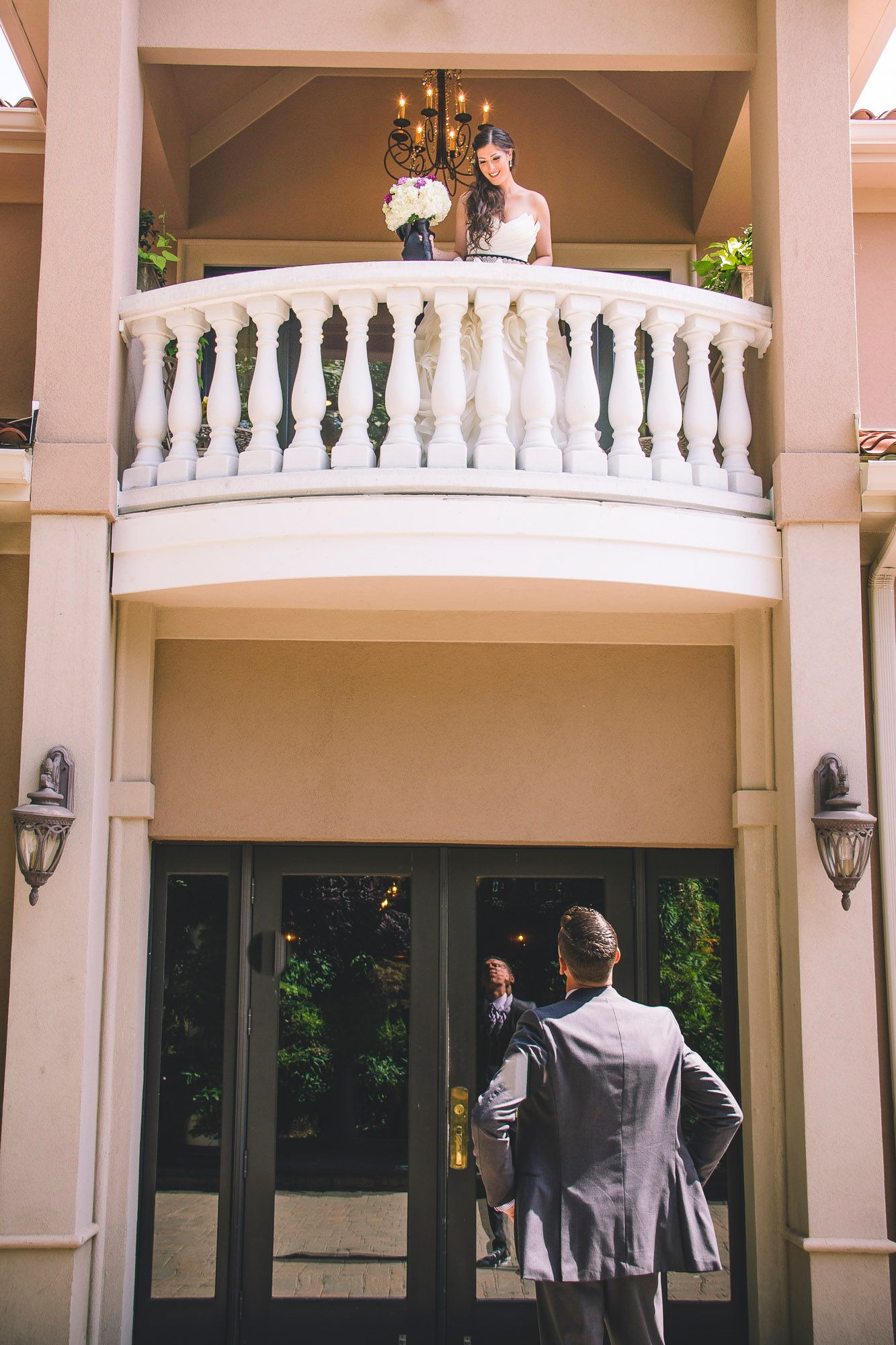 Bride stands on elegant balcony holding bouquet while groom looks up from below at venue entrance.