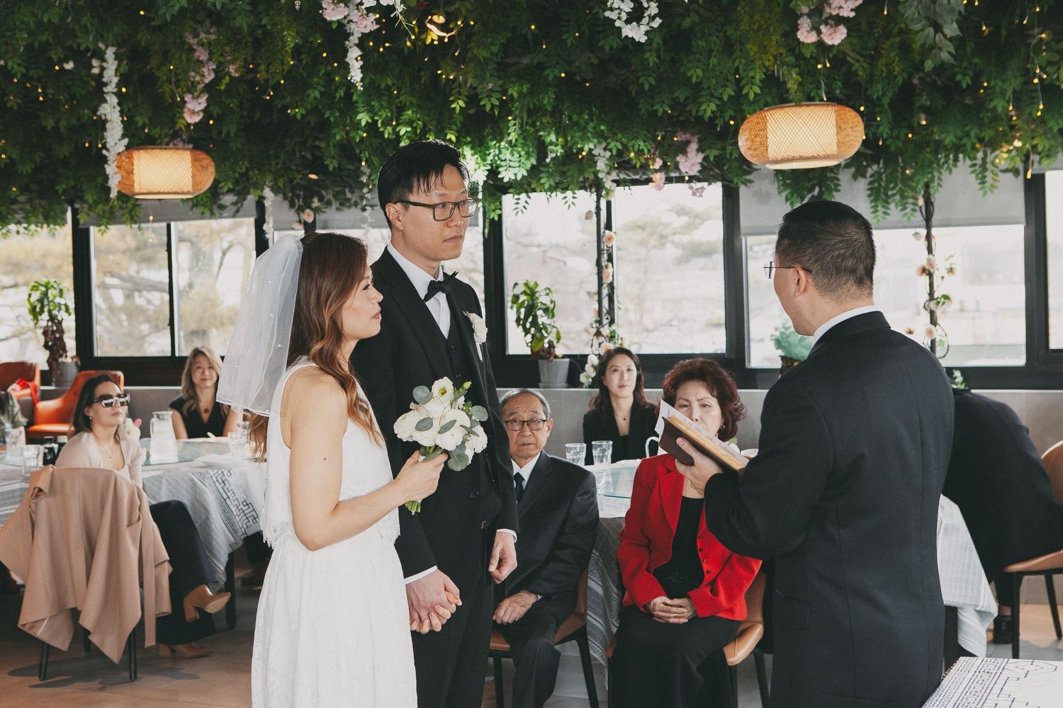 Asian bride and groom exchange vows at elegant indoor wedding ceremony with lush greenery and lantern decorations.