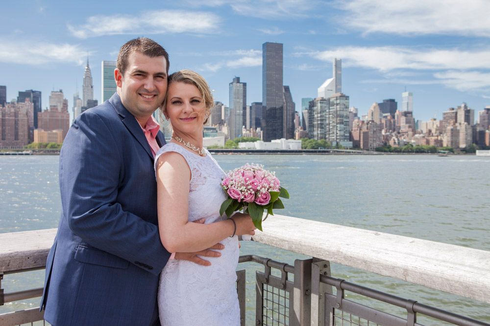 Smiling newlyweds pose together with Manhattan skyline and river in background, bride holding pink bouquet.