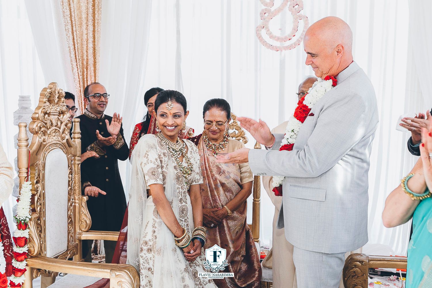 Bride in gold lehenga smiles beside tall groom in grey suit during Indian wedding ceremony.