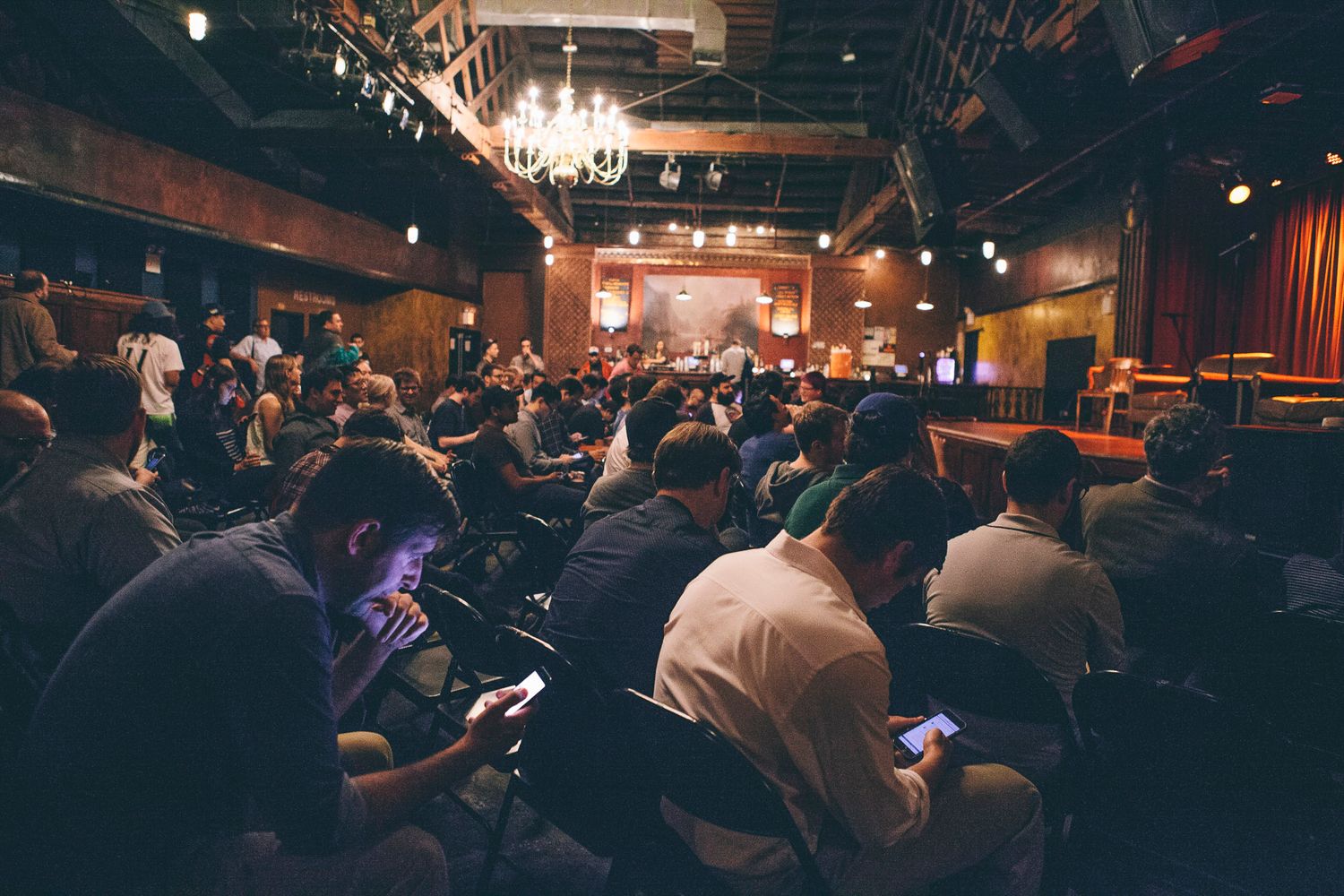 Crowd of attendees using smartphones during a live event in a dimly lit venue with stage.