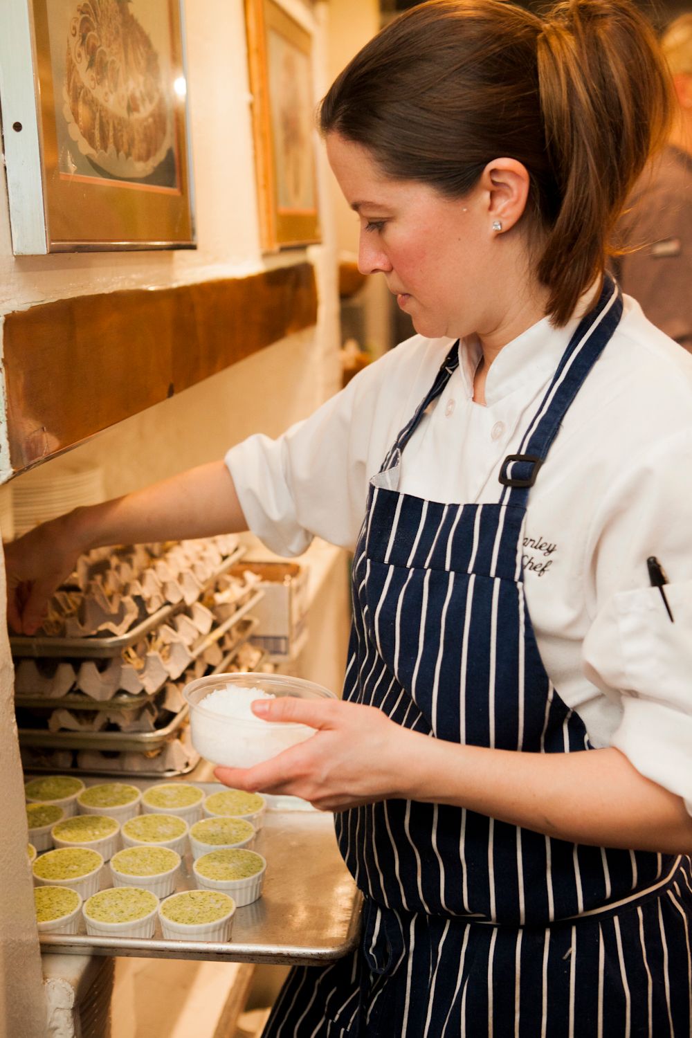 Female chef in striped apron preparing individual ramekins of food in a professional kitchen.