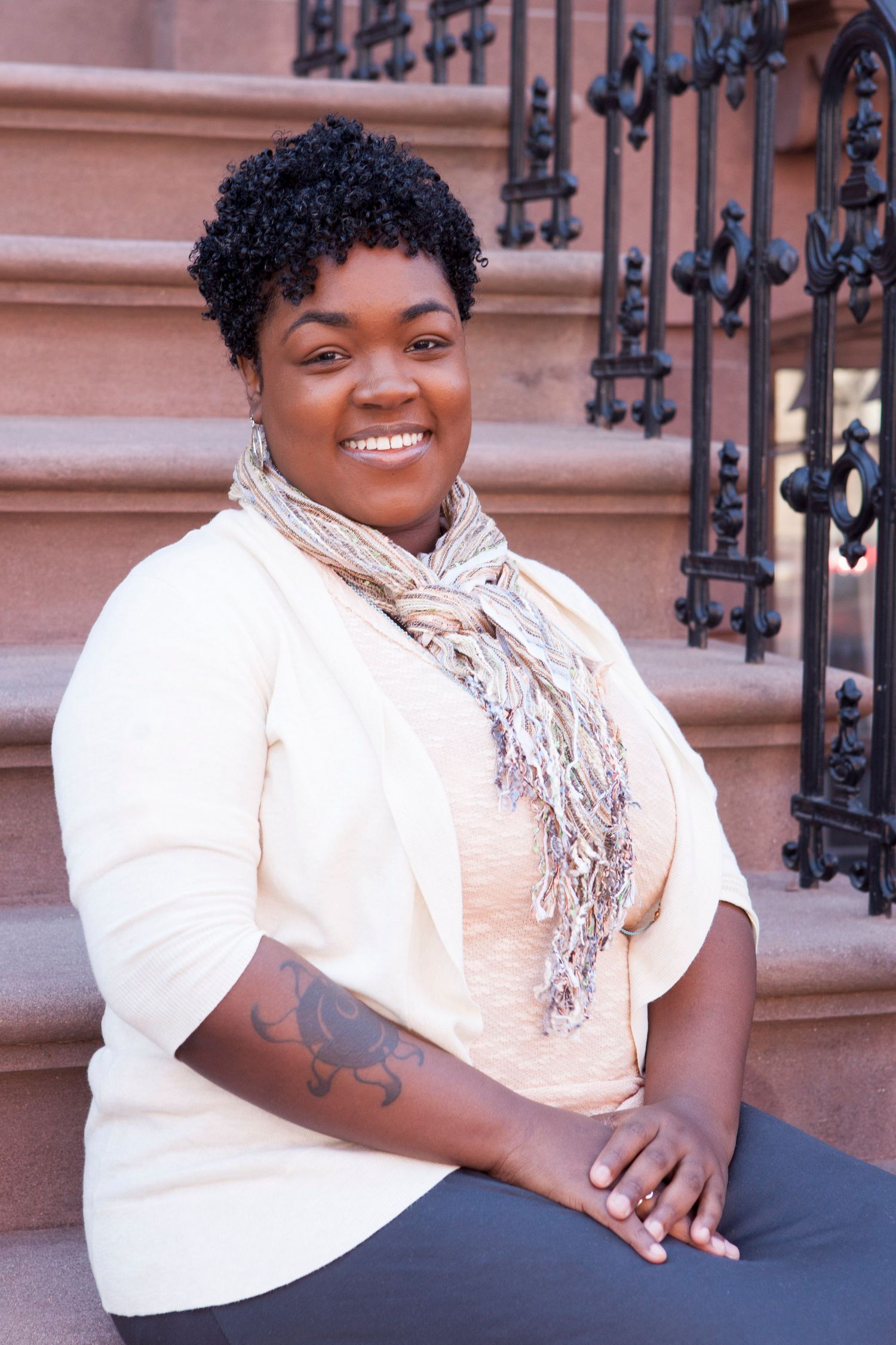 Smiling Black woman with short natural hair wearing a scarf, sitting on outdoor steps.