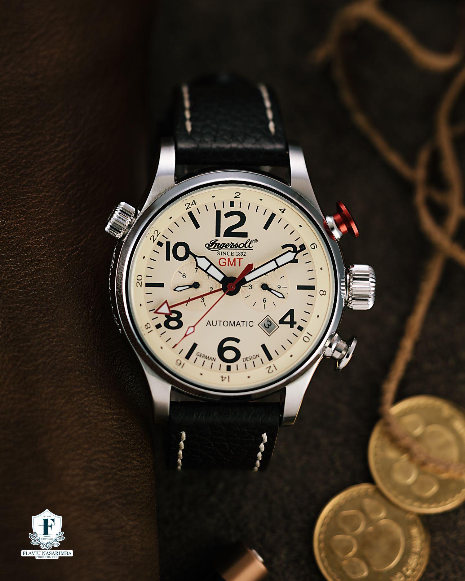Ingersoll GMT automatic watch with cream dial and black leather strap beside vintage coins.
