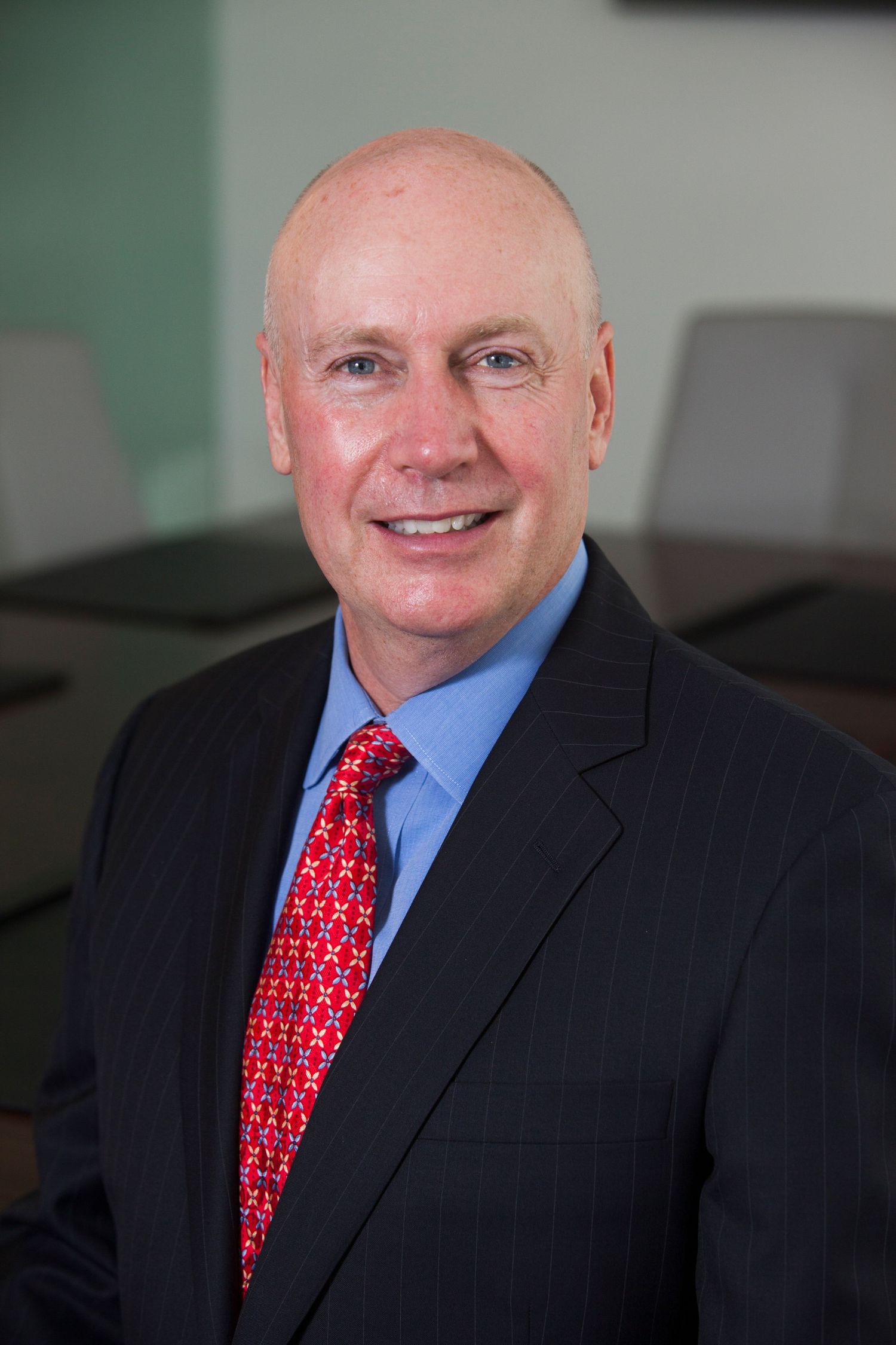 Smiling bald businessman in dark suit with red tie poses for professional headshot in modern conference room.