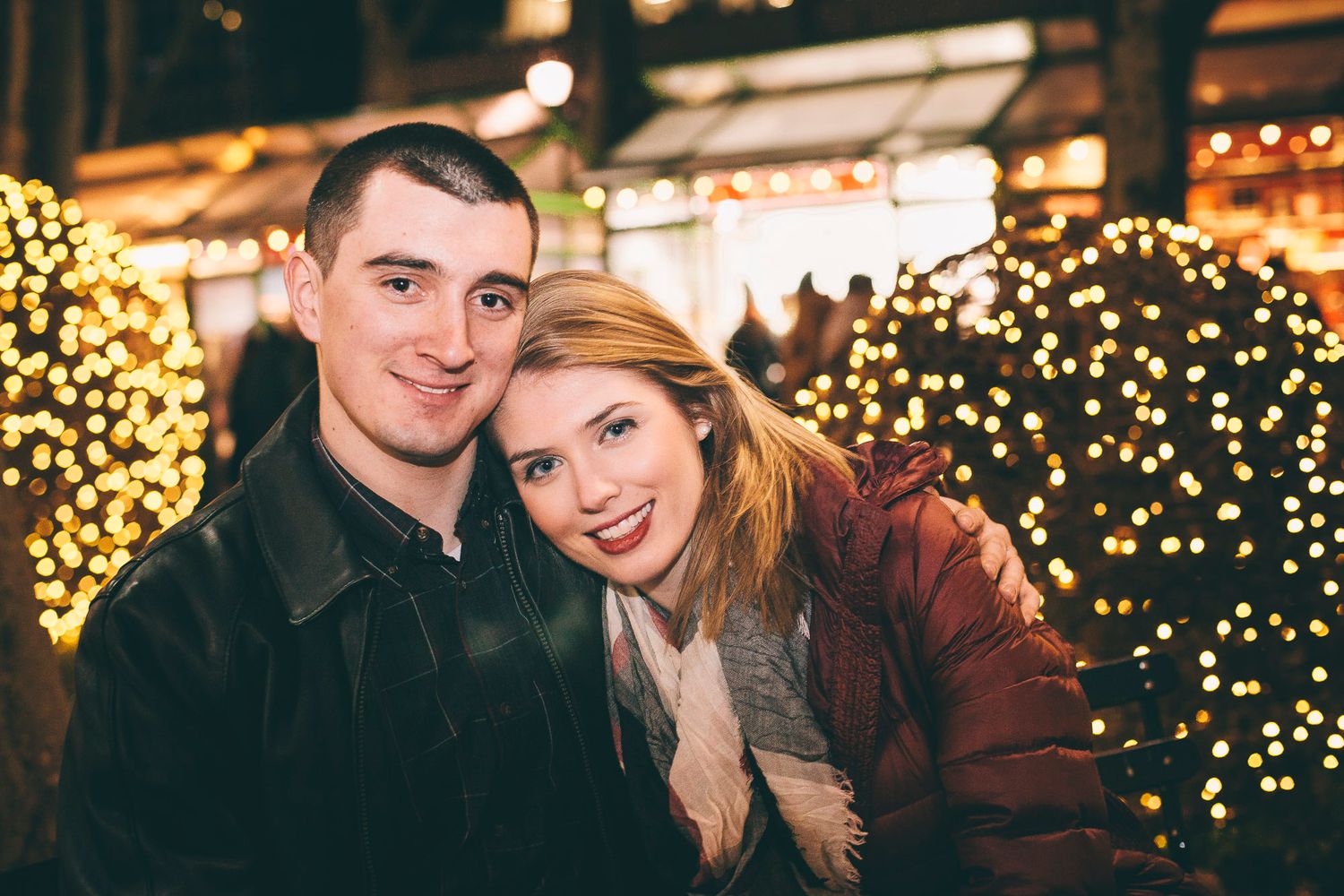 Smiling couple poses cheek to cheek surrounded by warm golden Christmas lights at night.
