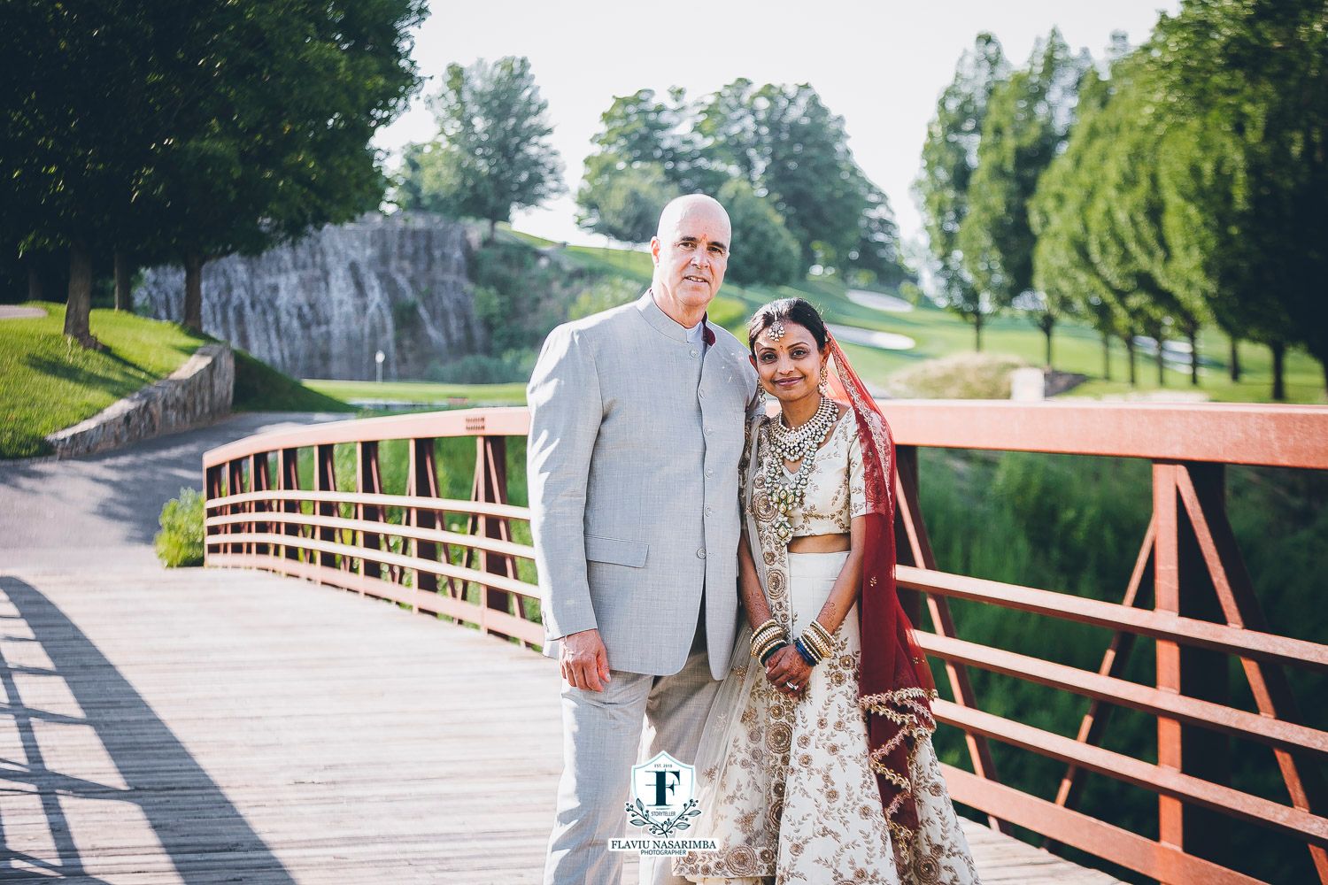 Interracial wedding couple in traditional Indian bridal attire posing on wooden bridge at golf course.