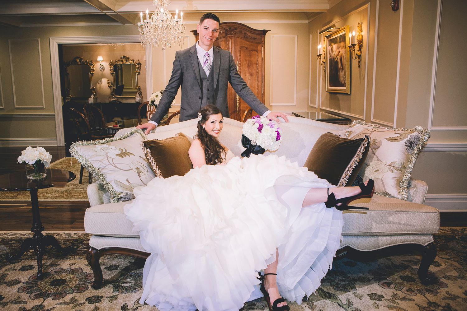 Bride in ruffled white gown reclines on ornate sofa while groom stands behind her at wedding.