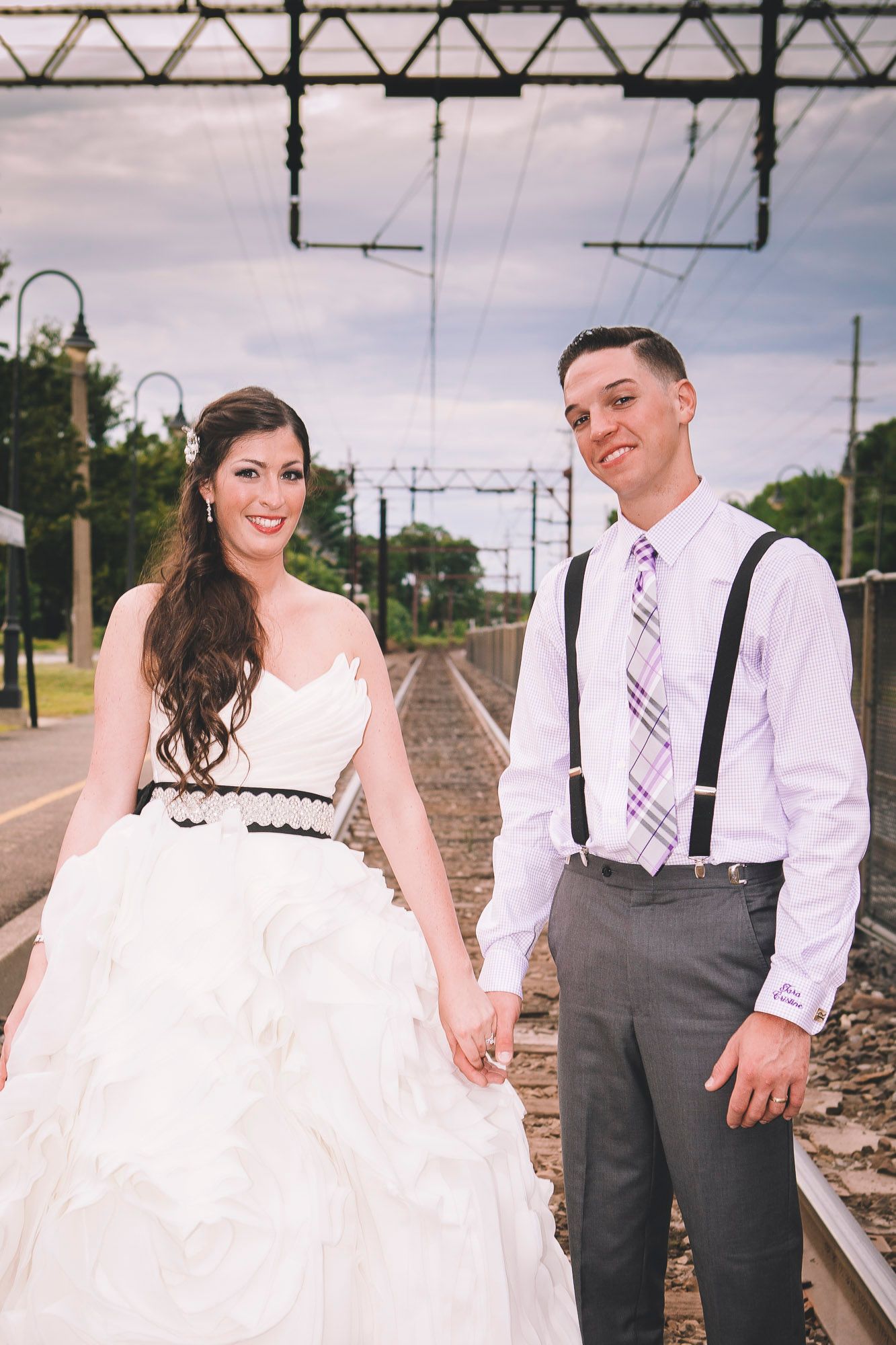 Bride and groom holding hands and posing together on railroad tracks in wedding day attire.