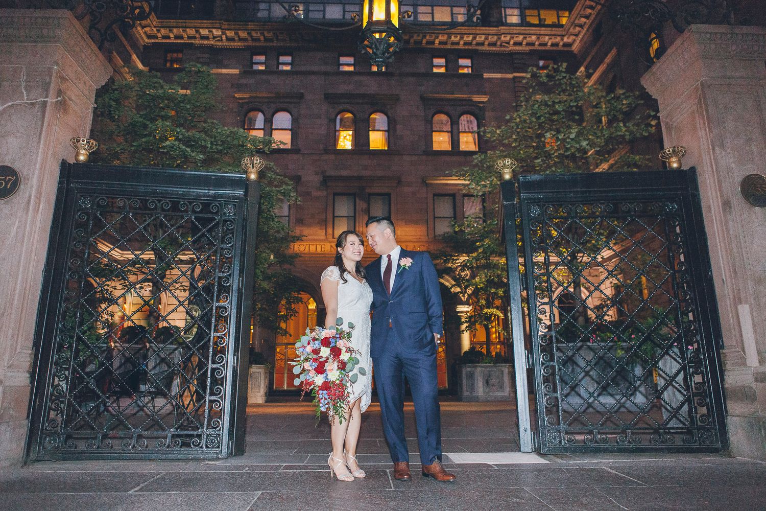 Bride and groom stand together at ornate iron gates outside a stunning lit building at night.