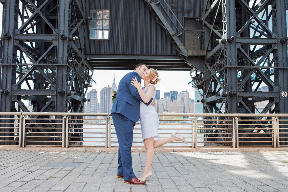 Newlyweds share a romantic kiss under large metal bridge structure with NYC skyline visible behind them.