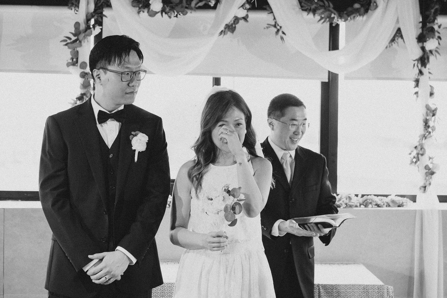 Emotional bride wipes tears during black and white wedding ceremony with groom and officiant.