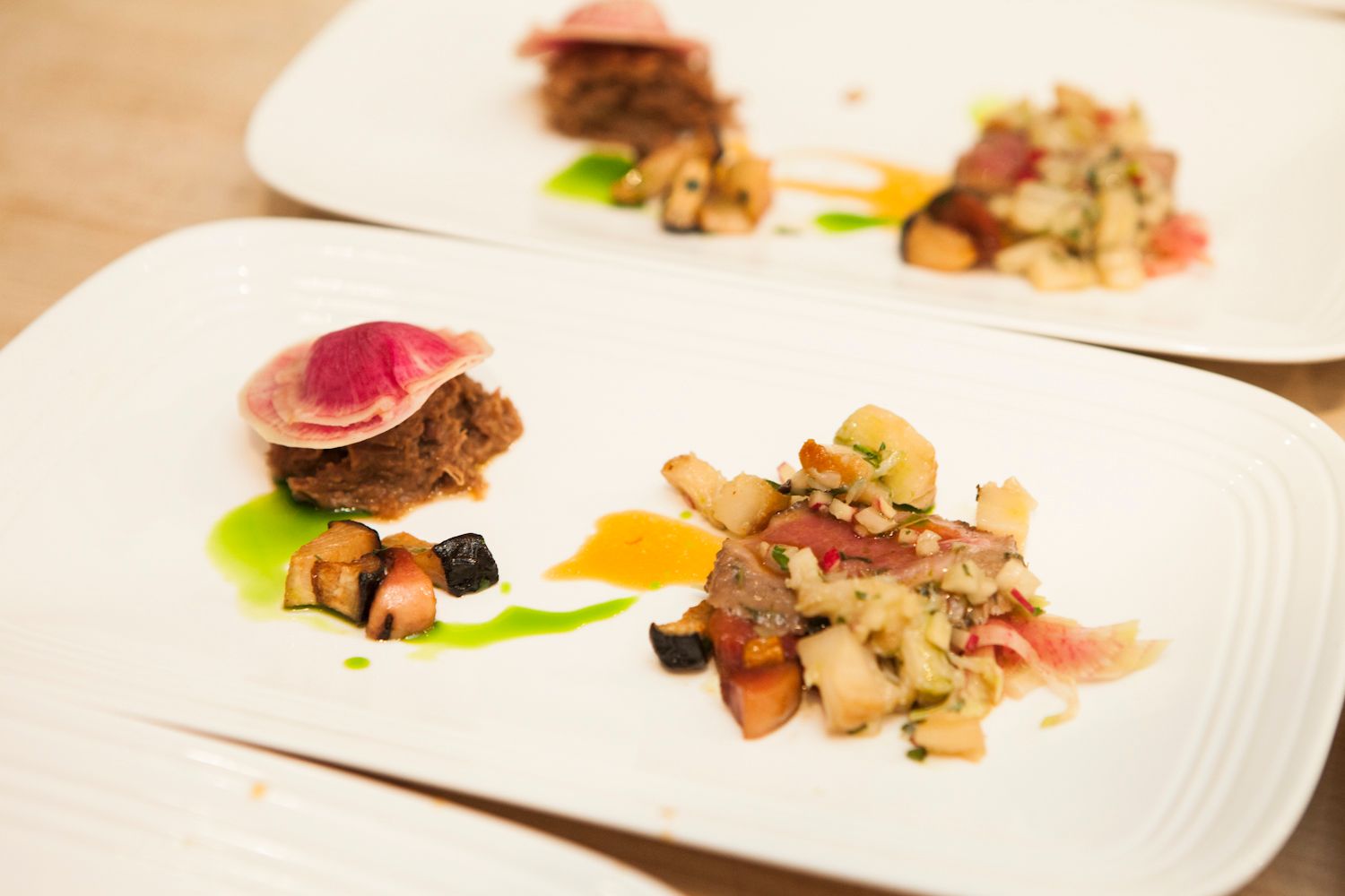 Gourmet plated dish with seared meat, colorful vegetables, and artistic sauce on white restaurant plates.