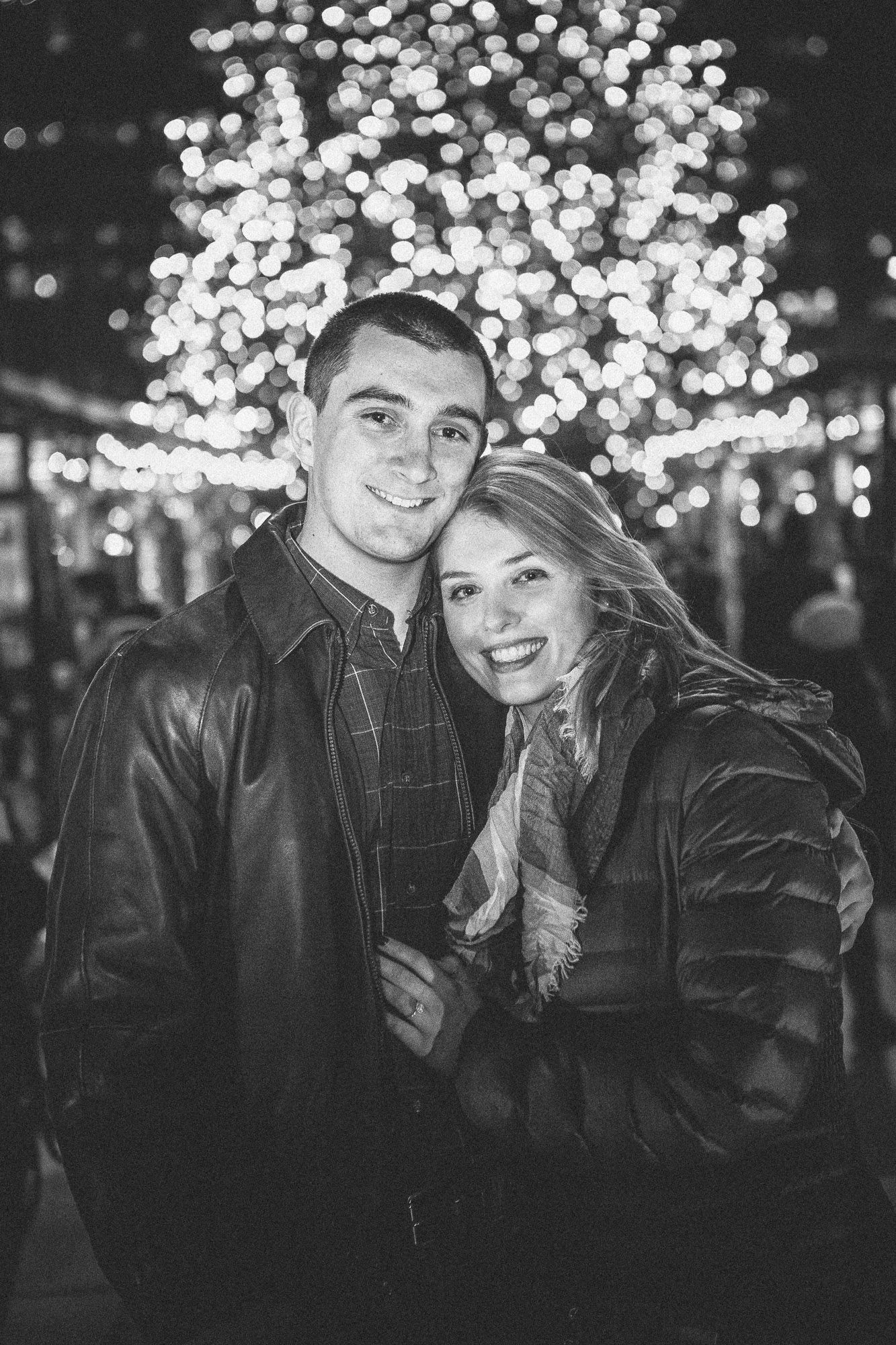 Black and white portrait of smiling couple cuddling before glowing Christmas tree at night.