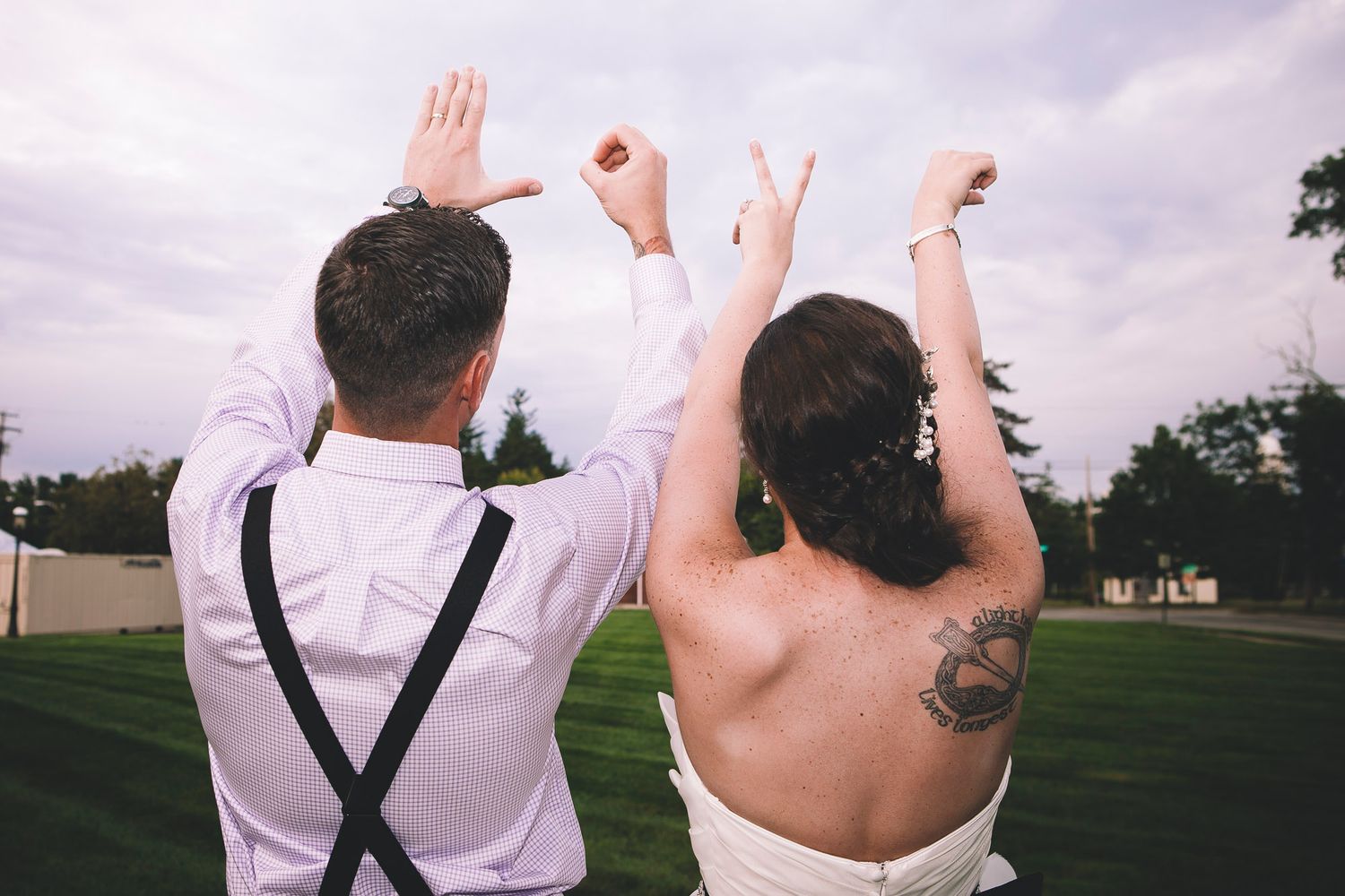 Bride and groom seen from behind raising hands outdoors, bride showing a decorative back tattoo.