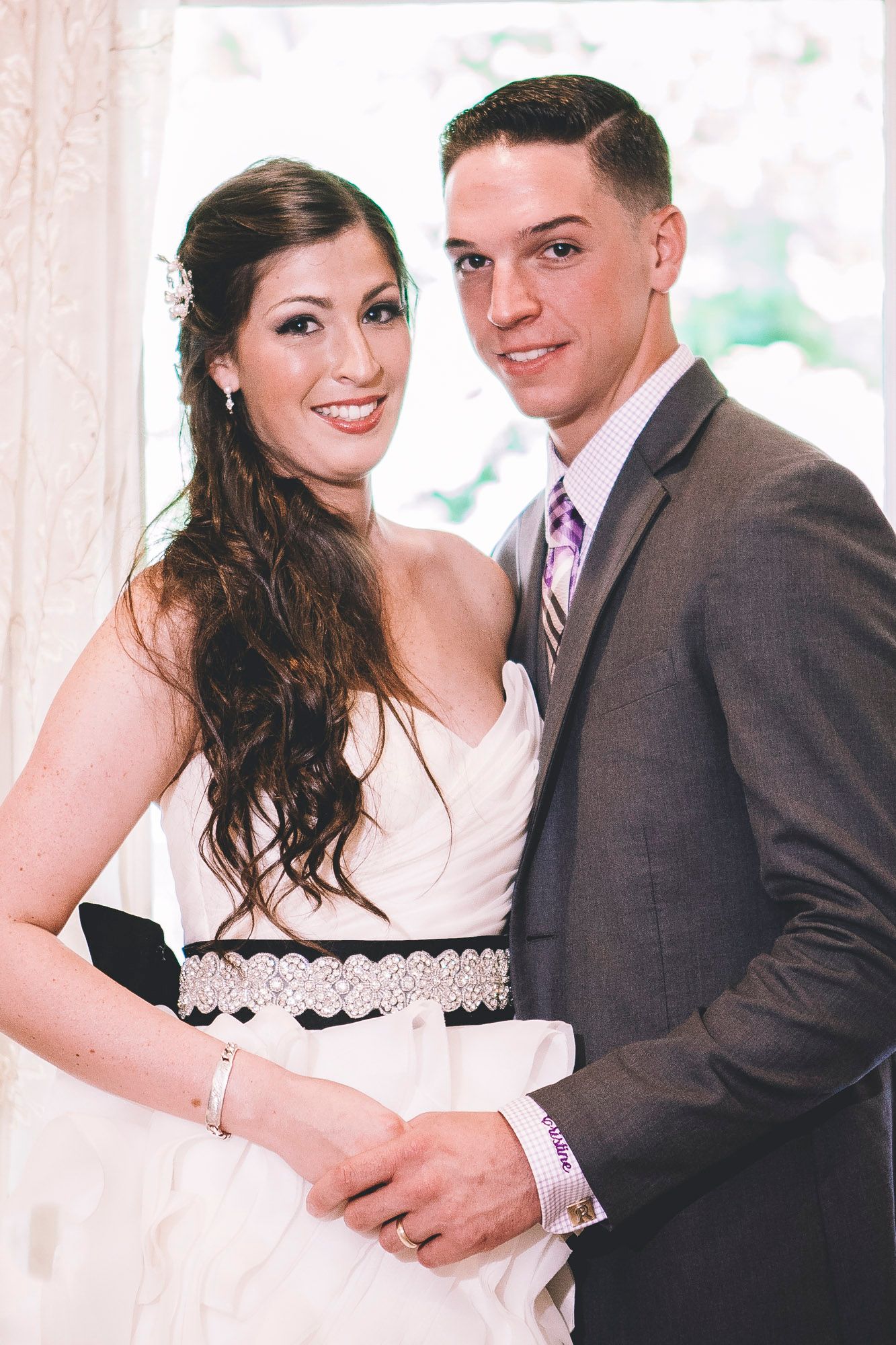 Smiling bride in white gown with black sash poses with groom in gray suit at wedding.