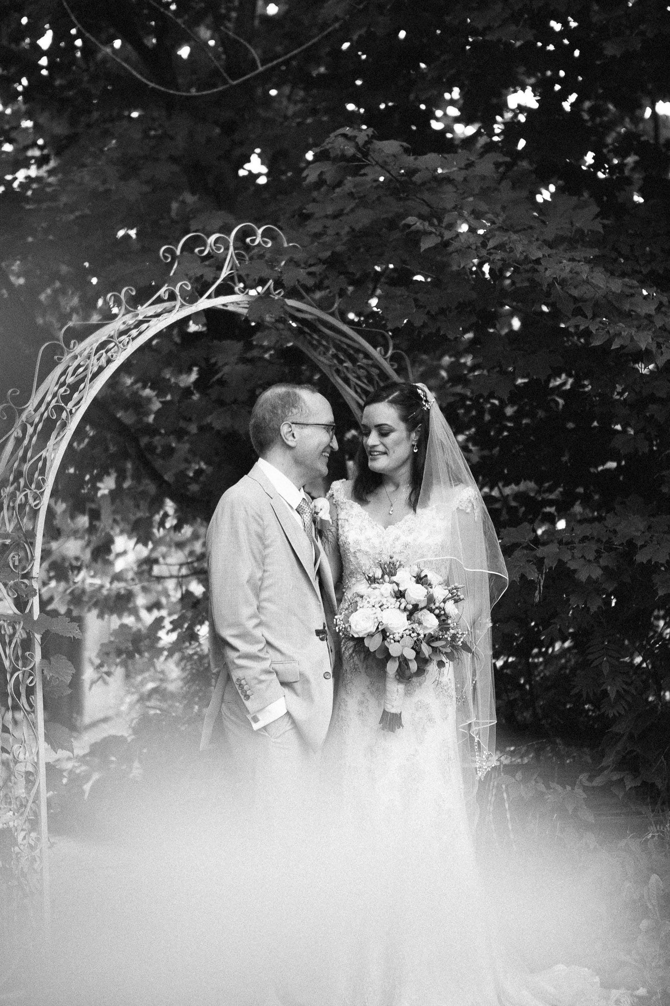 Black and white portrait of bride and groom sharing a tender moment under a garden arch.