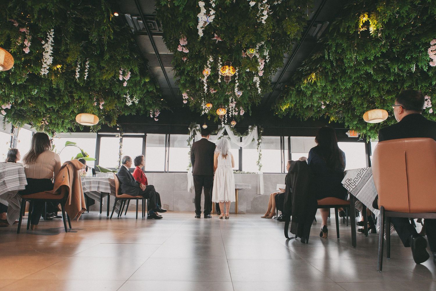 Wedding ceremony in a lush green venue with hanging plants as guests stand along the aisle.