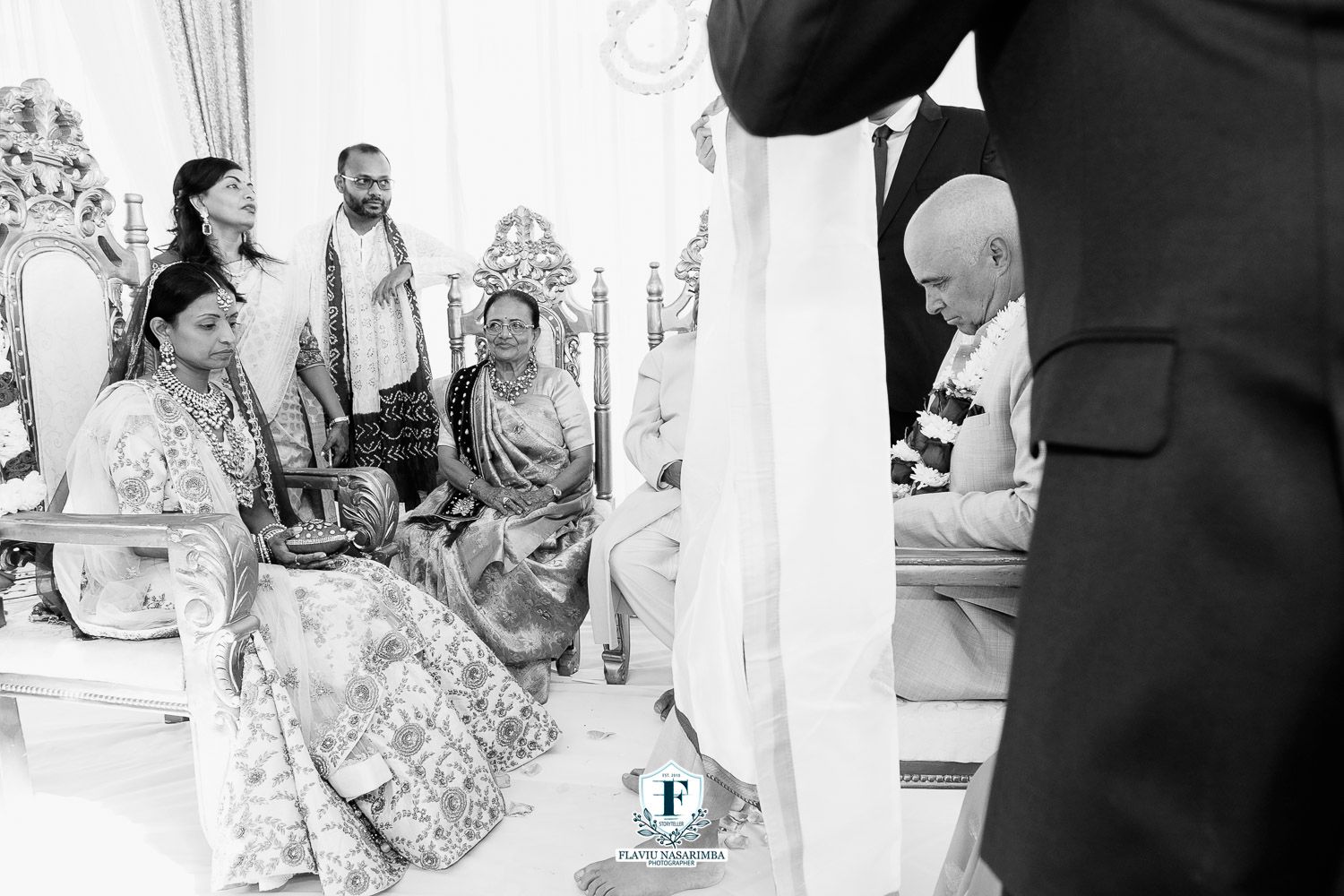 Family members in traditional Indian attire seated watching Hindu wedding ceremony at decorated mandap.