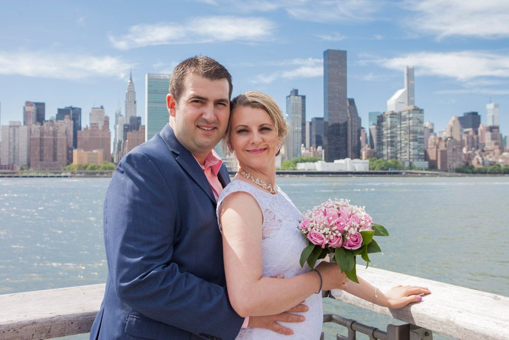 Couple embraces on NYC waterfront, groom in navy suit hugging bride holding pink rose bouquet.