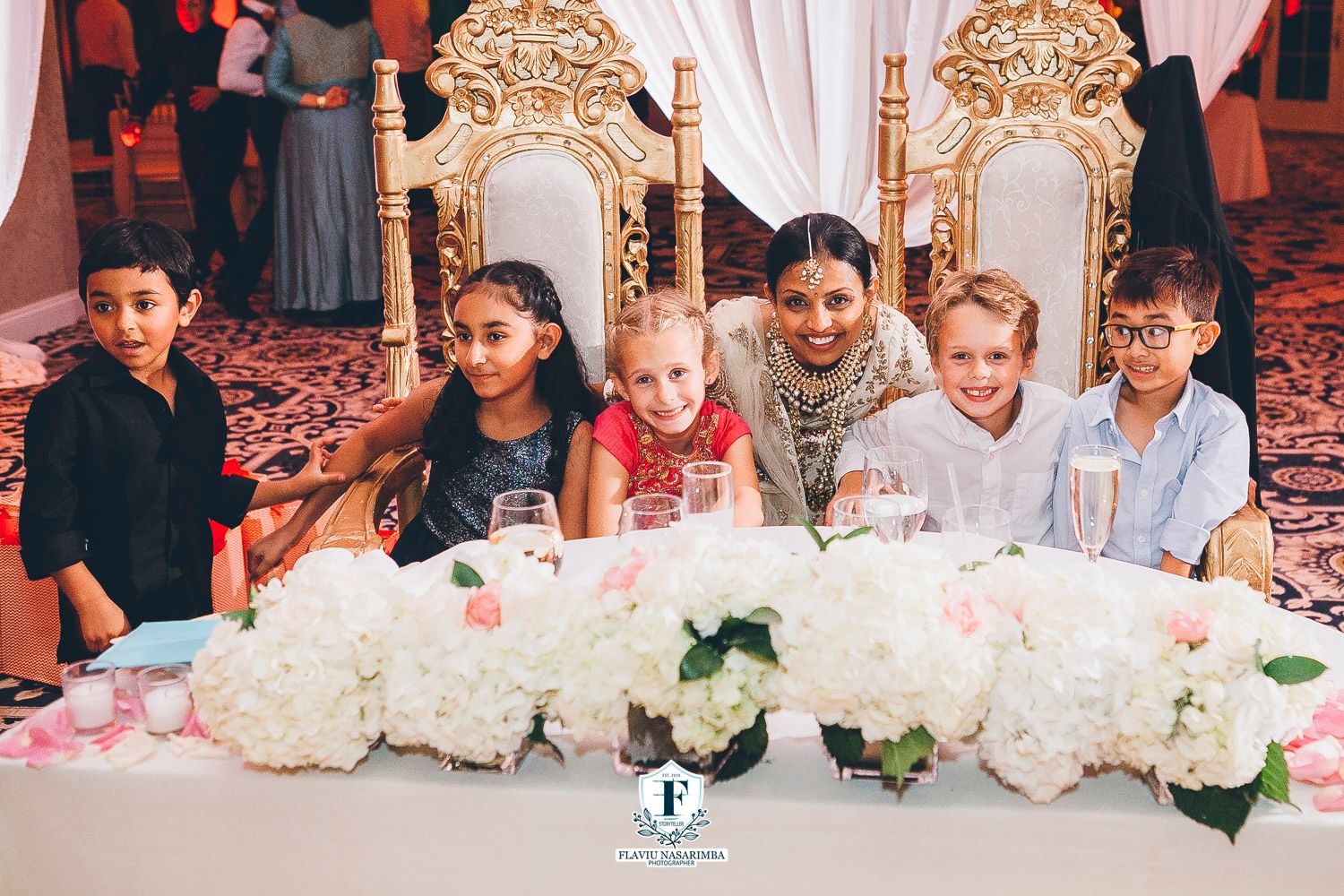 Children pose together at an elegant wedding reception with white floral arrangements and ornate chairs.