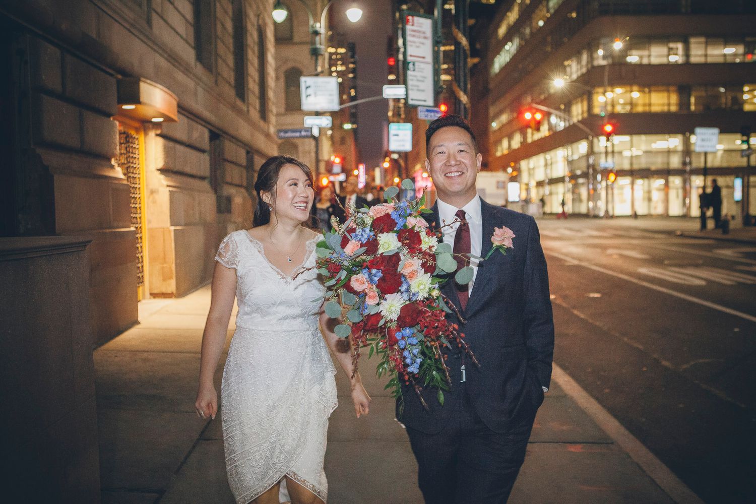 Bride and groom walking city streets at night, bride holding large red and blue bouquet in wedding attire.
