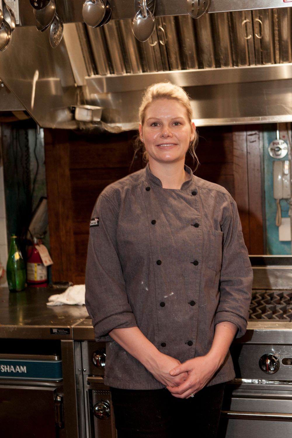 Female chef in gray double-breasted jacket standing confidently in a professional restaurant kitchen.