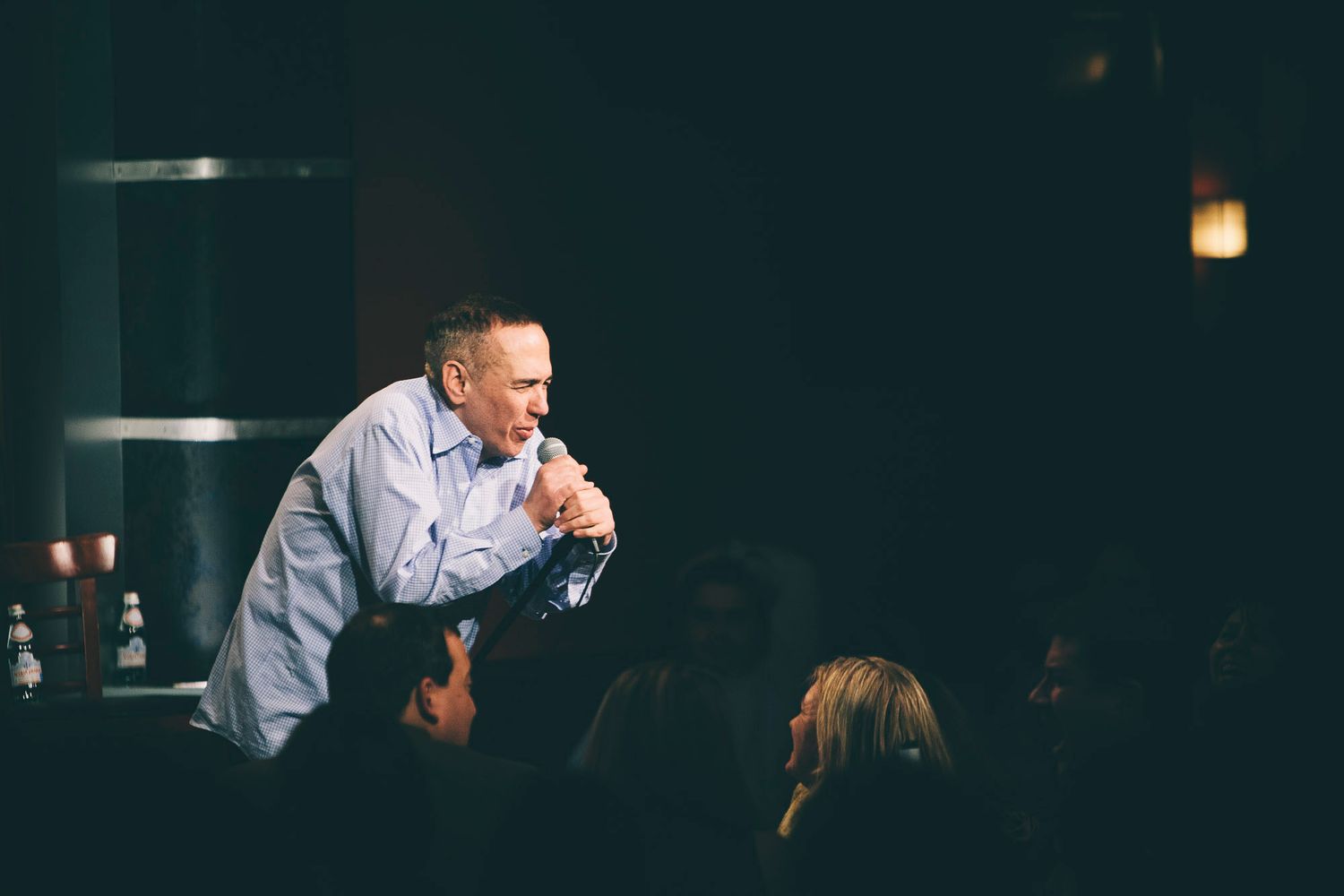 Male comedian leaning forward engaging with audience in dimly lit comedy club.
