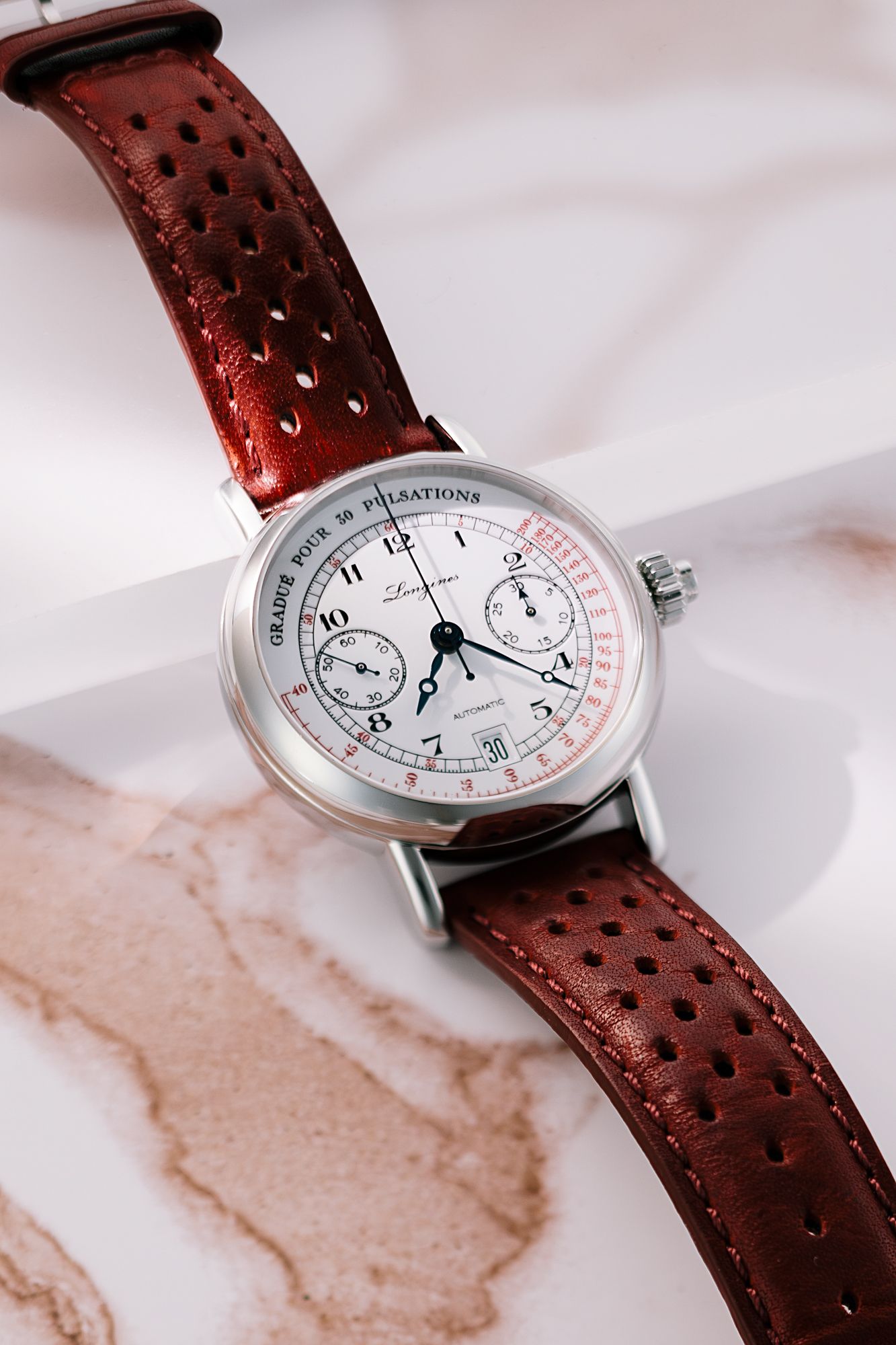 Elegant chronograph watch with white dial and burgundy perforated leather strap displayed on marble surface.