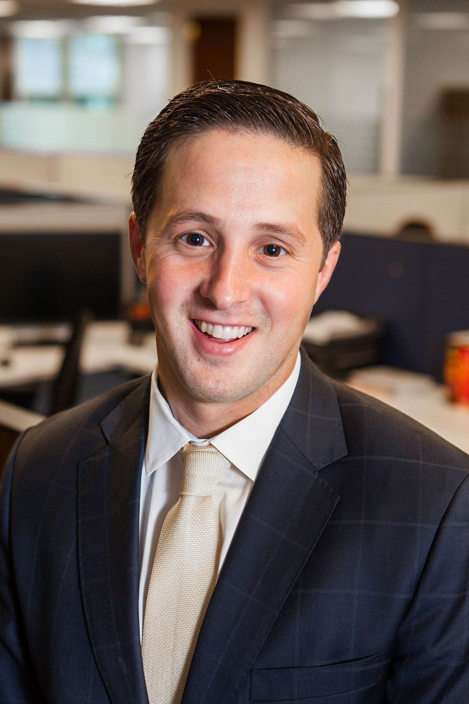 Smiling businessman in navy suit and tan tie posing in a modern office environment.