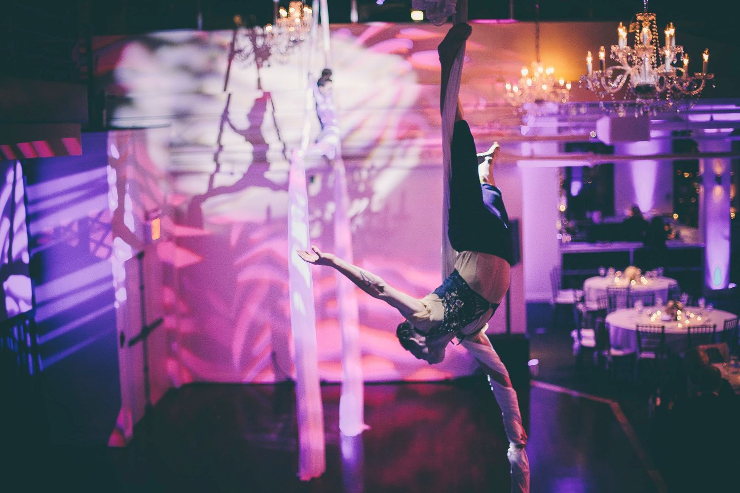 Aerial performer hanging upside down on silks beside a stilt walker in an elegant event venue.