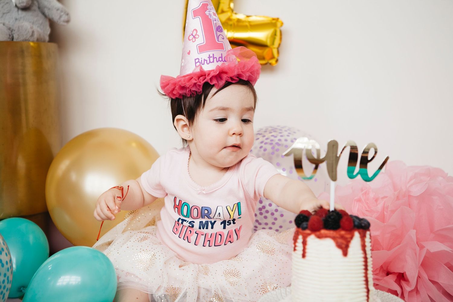 Baby girl wearing pink birthday hat and tutu reaches toward her first birthday cake with 'one' topper.