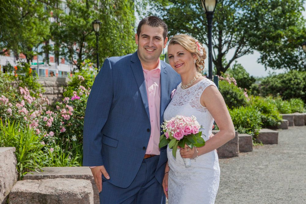 Happy couple poses together in garden, groom in navy suit and bride in white lace dress with pink bouquet.