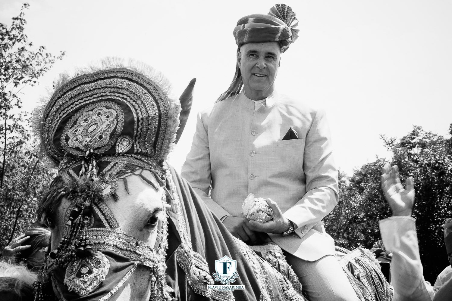 Groom in white sherwani and turban rides decorated horse at traditional Indian wedding baraat ceremony.
