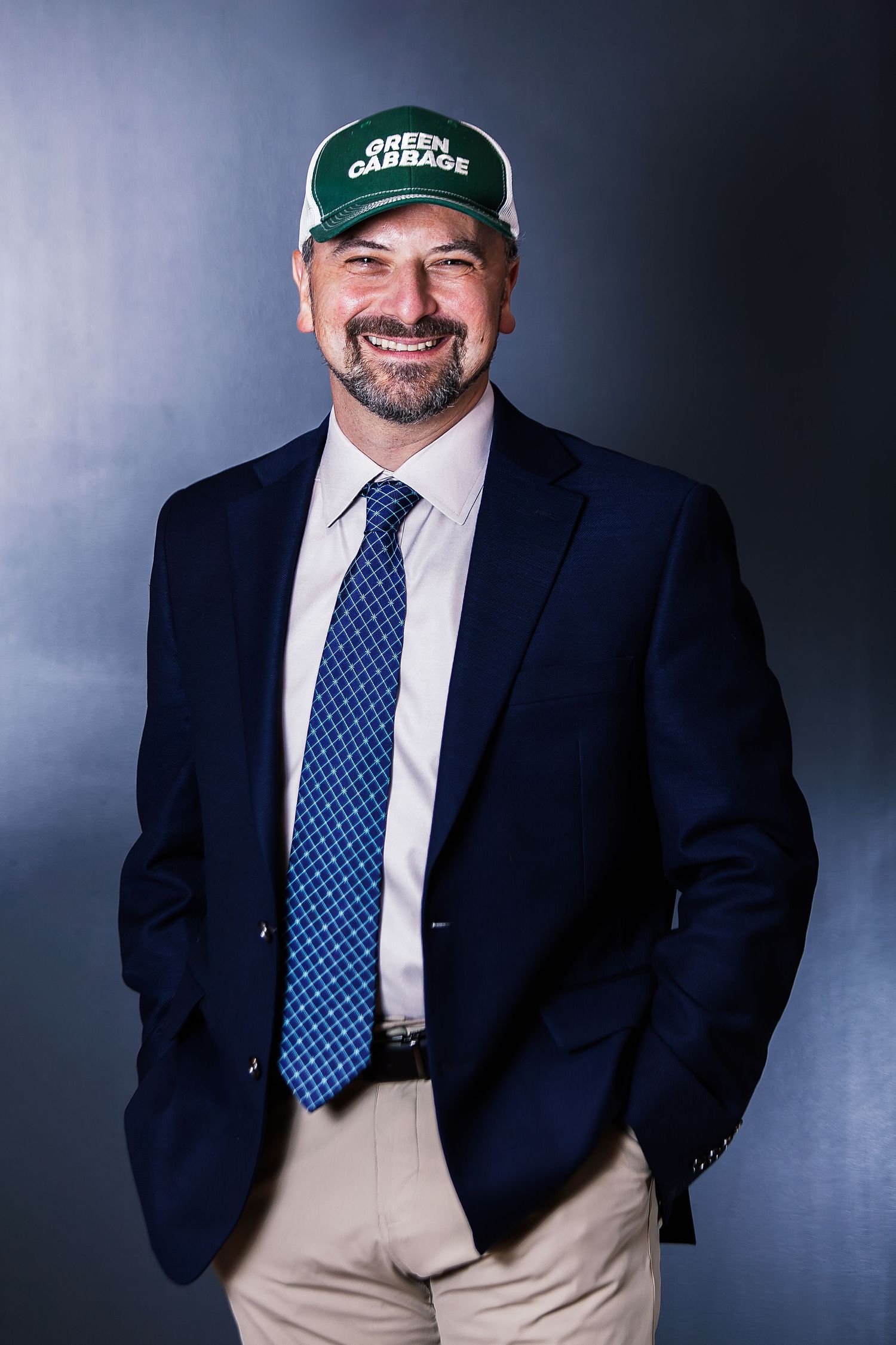 Man in navy blazer, blue tie, and green Green Garbage cap smiles confidently against a dark studio background.