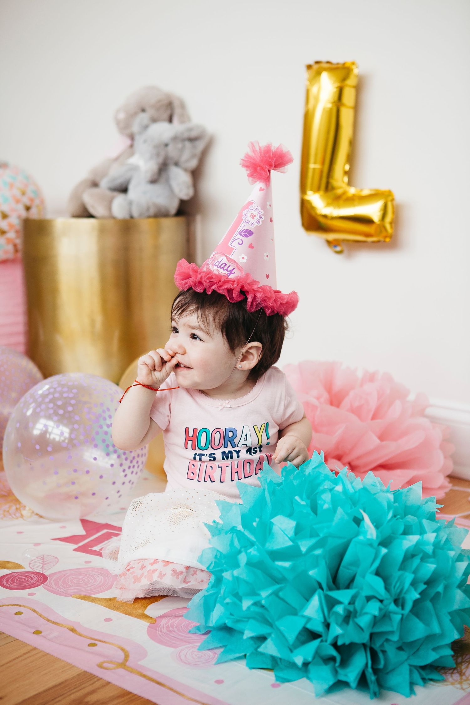 Adorable baby in pink party hat sits among teal pom poms and balloons celebrating her first birthday.